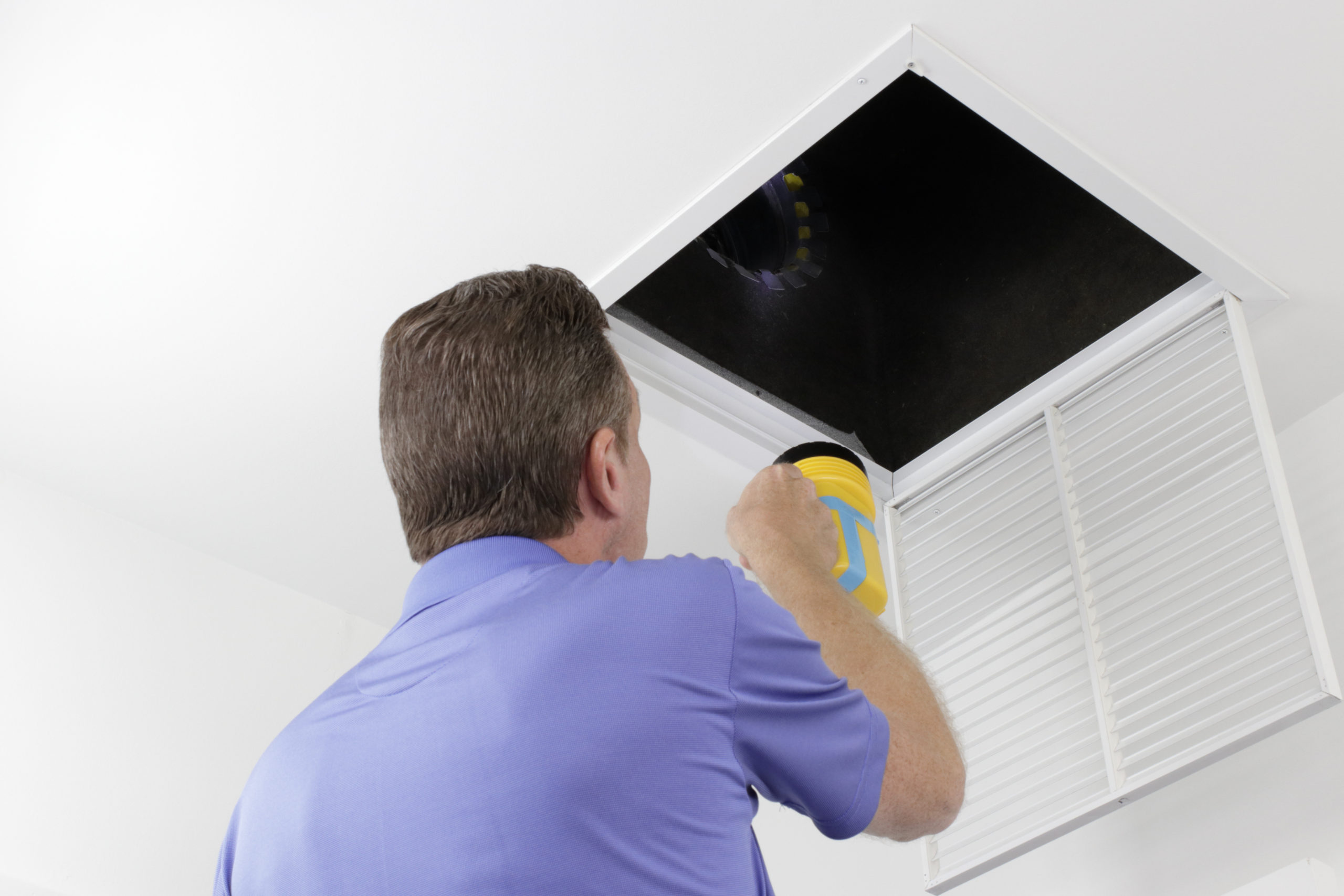 Older male with a yellow flashlight examining HVAC ducts in a large square vent. Male technician looking over the air ducts inside a home air intake vent.
